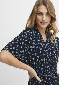 Marinblå klänning med korta ärmar och V-ringning, prydd med ett grönt blommönster, smal midja och mjuk tygtextur.