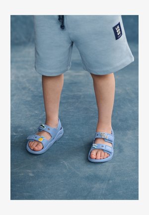 Šviesiai mėlynos basutės su dviem reguliuojamais dirželiais, puoštos spalvingais vaisių ir gyvūnų dekoracijomis. Minkšta medžiaga ir apvalus dizainas.