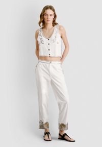 Bijela kratka majica s gumbima i srebrnim ukrasima, uparena s bijelim hlačama koje imaju dekorativne zlatne i srebrne obrube na rubu.