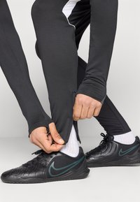 Μαύρο αθλητικό κολάν με εφαρμοστή σχεδίαση, που διαθέτει φερμουάρ στο πόδι και λευκή λεπτομέρεια στο πλάι. Ζευγάρι μαύρα αθλητικά παπούτσια.