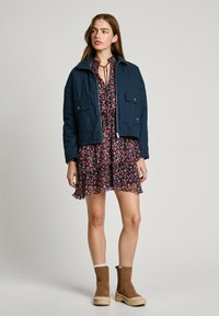 Giacca cropped blu navy con due tasche frontali, abbinata a un vestito nero con stampa floreale e volant, e stivali alla caviglia color tan con suole testurizzate.