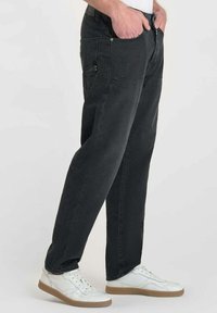 Pantalons en velours noir avec une coupe droite. Présente plusieurs poches et une fermeture éclair. Portés avec des baskets blanches.