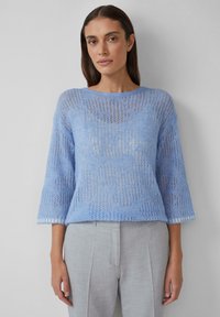 Hellblauer Strickpullover mit offenen Mustern, weiten Dreiviertelärmeln und Rundhalsausschnitt, kombiniert mit hellgrauen Hosen.