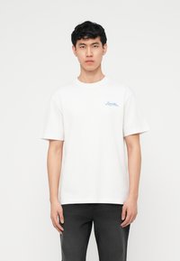 Νεαρός με  κοντά μαύρα μαλλιά, φοράει λευκό μπλουζάκι με μικρό μπλε κείμενο στο αριστερό μέρος του στήθους και μαύρο παντελόνι, στέκεται μπροστά σε λευκό φόντο.