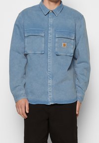 Camicia in denim azzurro chiaro con due tasche sul petto e un'etichetta circolare con logo. Maniche lunghe e colletto con chiusura a bottoni.