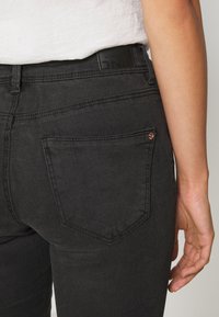 Zwarte spijkerbroek heeft een strak ontwerp met een achterzak en een gladde textuur. Een rechthoekig zwart label is zichtbaar aan de tailleband.