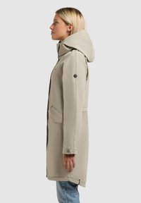 Femeie în picioare, purtând o haină bej cu glugă și blugi albaștri, cu fața spre stânga, pe un fundal simplu gri deschis.