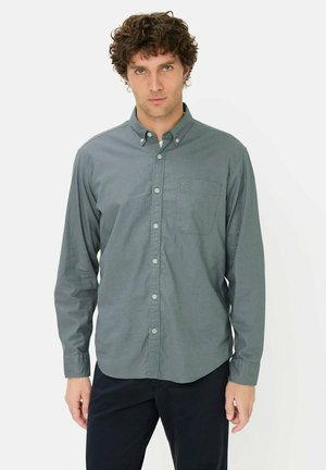 Mand med krøllet brunt hår iført langærmet grå skjorte med knapper og sorte bukser, stående foran en hvid baggrund.