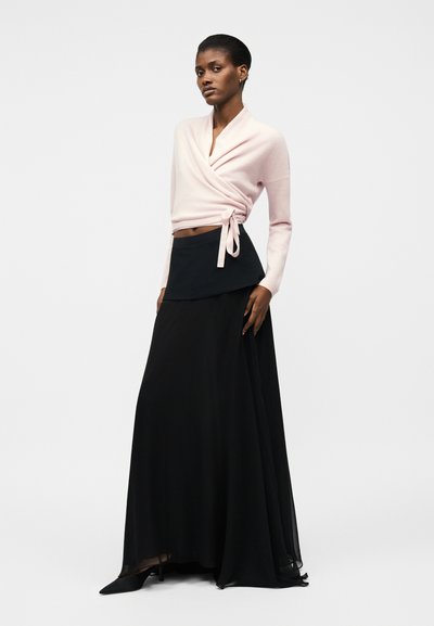 Kobieta w jasno-różowym swetrze kopertowym zawiązanym z boku oraz w zwiewnej czarnej maxi spódnicy i czarnych szpiczastych butach.