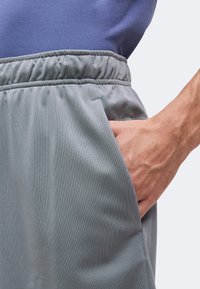 Main placée dans la poche latérale d'un pantalon gris à taille élastique porté avec une chemise bleue.