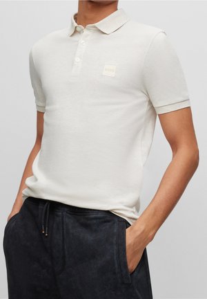 Lyse beige poloshirt med klassisk krave, tre-knaps lynlås og et lille firkantet logo-patch. Korte ærmer og blød tekstur.