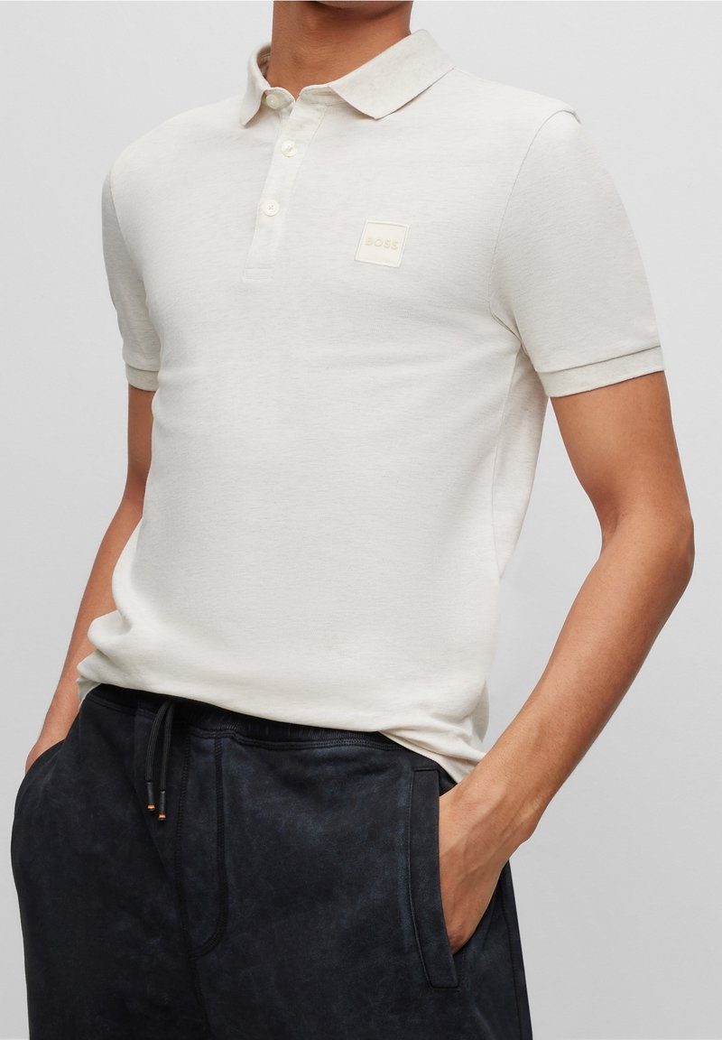 Polo beige clair avec un col classique, une patte de boutonnage à trois boutons et un petit patch logo carré. Manches courtes et texture douce.