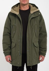 Chaqueta parka verde con capucha forrada de beige y suave, que presenta bolsillos con cremallera y botón, cordón ajustable y acentos de hardware visibles.