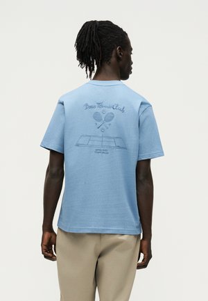 Άνδρας με dreadlocks που φοράει ένα ανοιχτό μπλε t-shirt «Boss Tennis Club» με γραφικά ρακέτες τένις και γήπεδο, μπεζ παντελόνι, γυρισμένος προς τα πίσω.