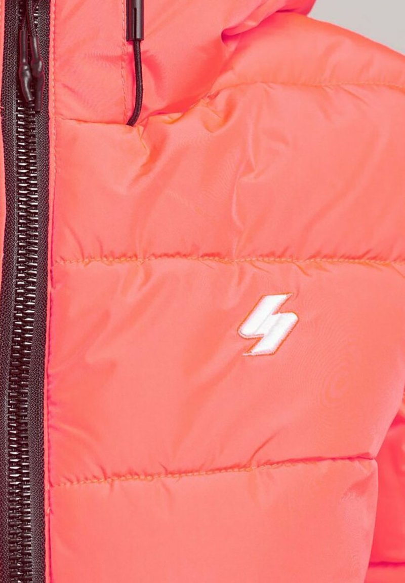 Primo piano di un giubbotto imbottito corallo con zip nera e piccolo logo a forma di fulmine bianco nella zona del petto.
