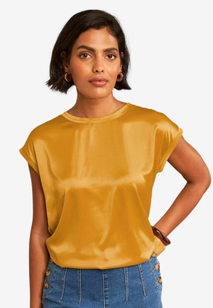 Kvinde med kort sort hår iført en sennepsgul satin top, blå knaplukket høj taljeskørt, brune øreringe i bøjleform og et armbånd.