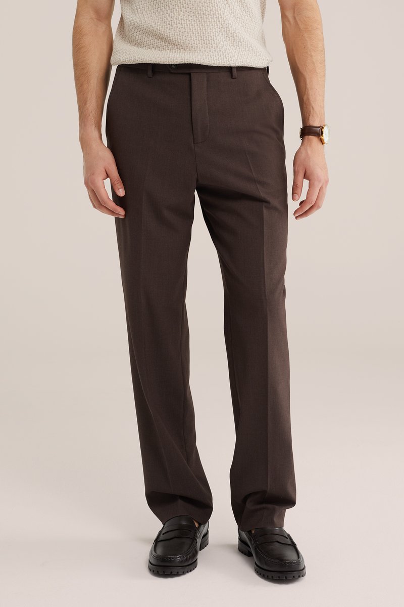 Homme portant un pantalon marron foncé taillé sur mesure, des mocassins noirs, un haut en maille crème et une montre avec un bracelet en cuir marron sur un fond neutre.
