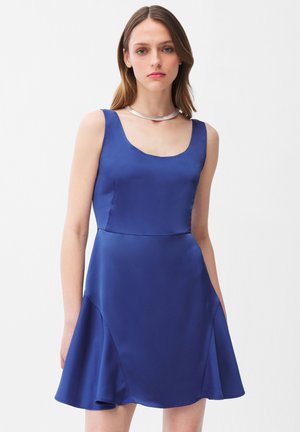 Młoda kobieta z włosami do ramion, ubrana w bezrękawnikową, królewską niebieską satynową sukienkę i srebrny naszyjnik, na białym tle.