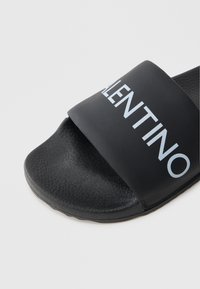 Czarne klapki na płaskiej podeszwie z szerokim paskiem, na którym znajduje się biały napis "VALENTINO". Teksturowana podeszwa z gładkim materiałem wierzchnim.