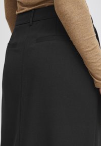 Jupe noire en tissu texturé, présentant une taille ajustée et de subtiles poches arrière. Un haut à manches longues de couleur beige complémentaire est visible.