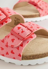 Vaaleanpunaiset sandaalit, joissa on nappa yläosa, jossa on punaisia sydänkuvioita, säädettävät punaiset soljet ja vaaleanruskea pehmustettu pohja valkoisella ulkopohjalla.