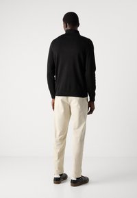 Pulover negru tricotat cu guler înalt, asortat cu pantaloni de culoare deschisă și pantofi negri. Textură netedă, design simplu, detalii minime.