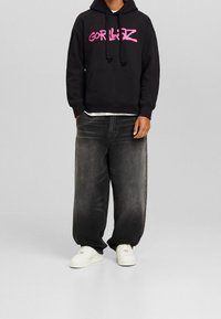 Svart huvtröja med rosa "GORLIZ"-text, framficka, dragsko på huvan, kombinerad med löst sittande mörkgrå jeans och vita sneakers.