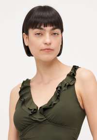 Olīvu zaļš peldkostīms ar vilnīšiem ap kakla un plecu līniju; mīksts, elastīgs audums ar pieguļošu siluetu.