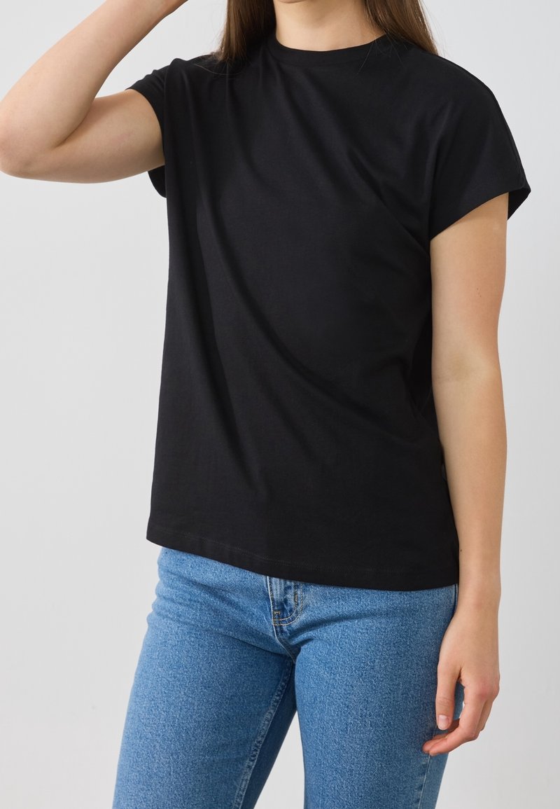 Personne portant un t-shirt noir à manches courtes et un jean bleu, avec un bras levé près de la tête sur un fond uni.