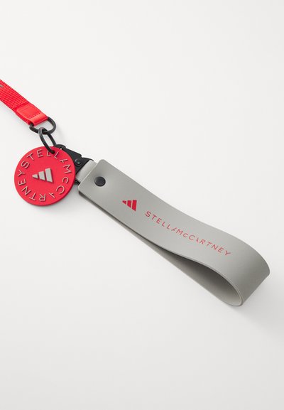 Harmaa silikoninen avaimenperä punaisella logotagilla, jossa on metallinen klipsi ja kohokuvioitu teksti "STELLA McCARTNEY." Sujuva pinta, moderni muotoilu.