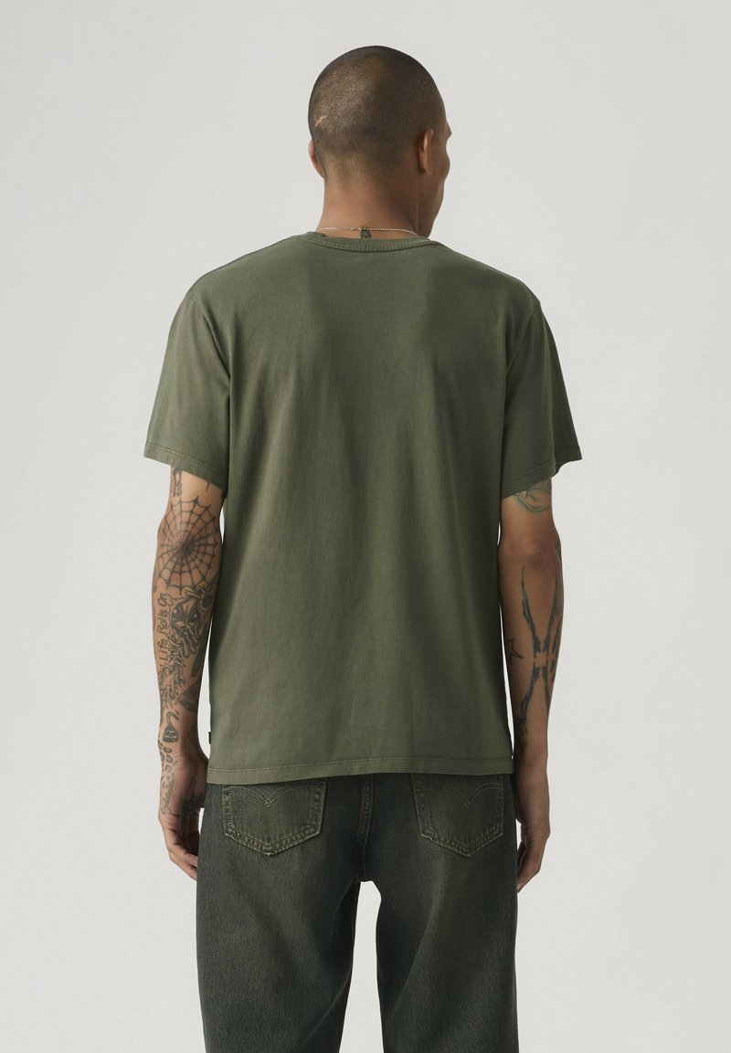 Man met kort haar, gekleed in een olijfgroen T-shirt en donkere spijkerbroek, toont tatoeages op beide armen, gezichten van zich afgewend tegen een effen achtergrond.