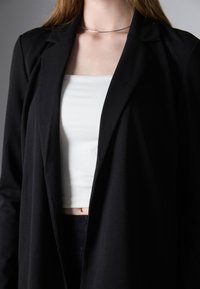 Kobieta ubrana w czarny żakiet na białą bluzkę bez ramiączek, z srebrnym naszyjnikiem choker, prezentująca się od ramion do talii na szarym tle.