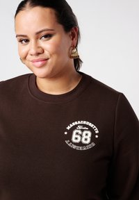 Bruine trui met ronde hals, voorzien van witte geprinte tekst en cijfers op de borst. De stof oogt zacht en glad. Statement oorbellen zichtbaar.