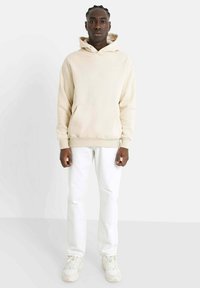 Sudadera de algodón beige con un bolsillo delantero y capucha con cordón, combinada con jeans de mezclilla blancos y zapatillas ligeras, de pie frente a un fondo neutro.