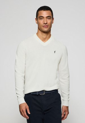 Witte v-hals trui van zachte stof, met lange mouwen en een klein zwart logo op de linkerborst. Gecombineerd met donkere broek en een geweven riem.