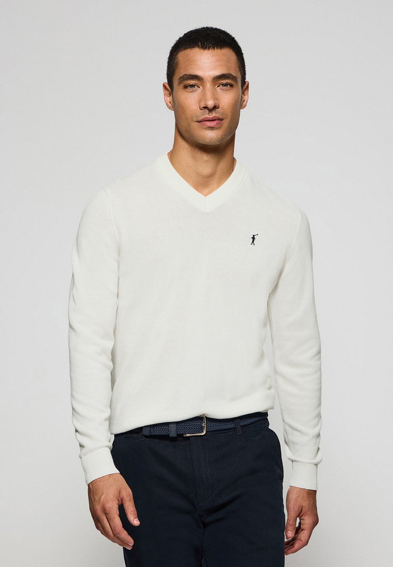 Weißer V-Ausschnitt-Pullover aus weicher Stoff, mit langen Ärmeln und einem kleinen schwarzen Logo auf der linken Brust. Kombiniert mit dunklen Hosen und einem gewebten Gürtel.