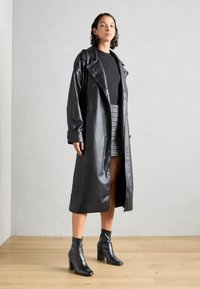 Trench coat de cuero sintético negro con botones de doblebreasted, amplias solapas y mangas largas. Combinado con una blusa negra y una falda estampada.