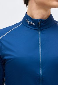 Primer plano de una persona con una chaqueta azul con cremallera y el logo "Rapha" en el cuello, con costuras reflectantes a lo largo del hombro.