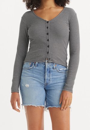 Femme portant un haut à manches longues rayé noir et blanc avec boutons, associé à un short en jean bleu clair usé avec ourlets effilochés.