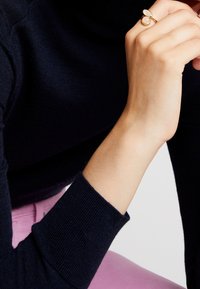 Guld ring med två ovala, blekrosa stenar. Buren på en hand med ljus hudton, som vilar mot en mörk tröja.