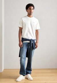 Λευκή μπλούζα με λαιμόκοψη, μπλε τζιν και λευκά αθλητικά παπούτσια. Μπλε ριγέ πλεκτό ύφασμα δεμένο στη μέση. Λείο, casual σχέδιο.