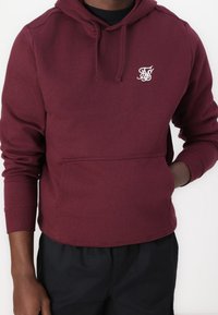 Uomo che indossa un hoodie color porpora con tasca frontale e logo ricamato sul petto, abbinato a pantaloni neri.