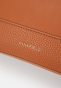 Tan leather handbag with textured surface and smooth accents. Features embossed logo "ANNA FIELD" in gold, positioned near seam.