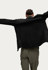 Chaqueta negra de gran tamaño hecha de una tela suave, con mangas largas y un dobladillo curvado. La parte trasera presenta un acabado liso y texturizado.