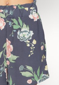 Detailní záběr na námořnické modré šortky s růžovým, žlutým a zeleným květinovým vzorem a elastickým pasem na pase osoby.