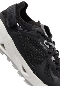 Schwarzer Sportschuh mit einem Mesh-Obermaterial und Perforationen, akzentuiert durch eine weiße Sohle und textured Details. Logo an der Ferse.