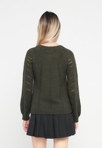 Oliwkowy sweter dzianinowy z teksturowanym wzorem, z długimi bufkami i pionowymi otwartymi ściegami, oraz ściągaczem na dole. Połączony z plisowaną czarną spódnicą.