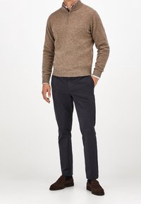 Brauner Strickpullover mit halbem Reißverschlusskragen, kombiniert mit dunkelblauen Slim-Fit-Hosen und braunen Wildlederschuhen. Glatte Texturen, lässiges Design.