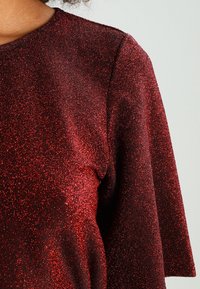 Top brillante rojo con escote redondo y mangas cortas. La tela tiene un acabado texturizado y brillante, resaltando un tono rico y profundo.