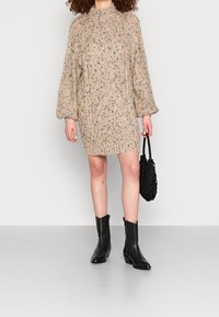 Beiges, gestricktes Sweatkleid mit einem strukturierten Muster und langen Ärmeln, kombiniert mit schwarzen Stiefeletten und einer schwarzen, geflochtenen Handtasche.